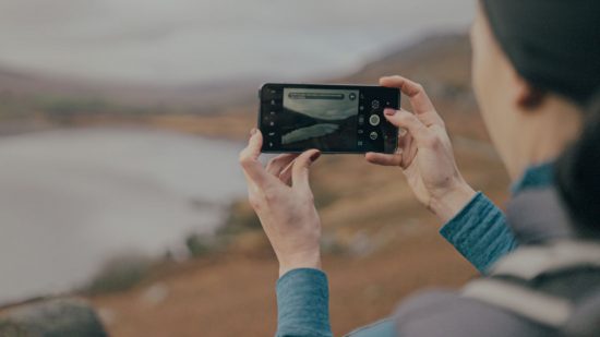 最高の頑丈な携帯電話の 1 つである Cat S75 は、丘陵地帯で写真を撮っている湖の方を向いている人の手に黒い携帯電話です。