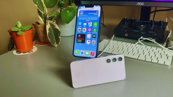 iPhone と Android のヘッダーには、iPhone 14 が黒、Samsung Galaxy S23 がラベンダーで表示されています。  iPhone はスタンドの上に置かれ、Samsung はその横に横たわり、背景には植物とキーボードが置かれています。