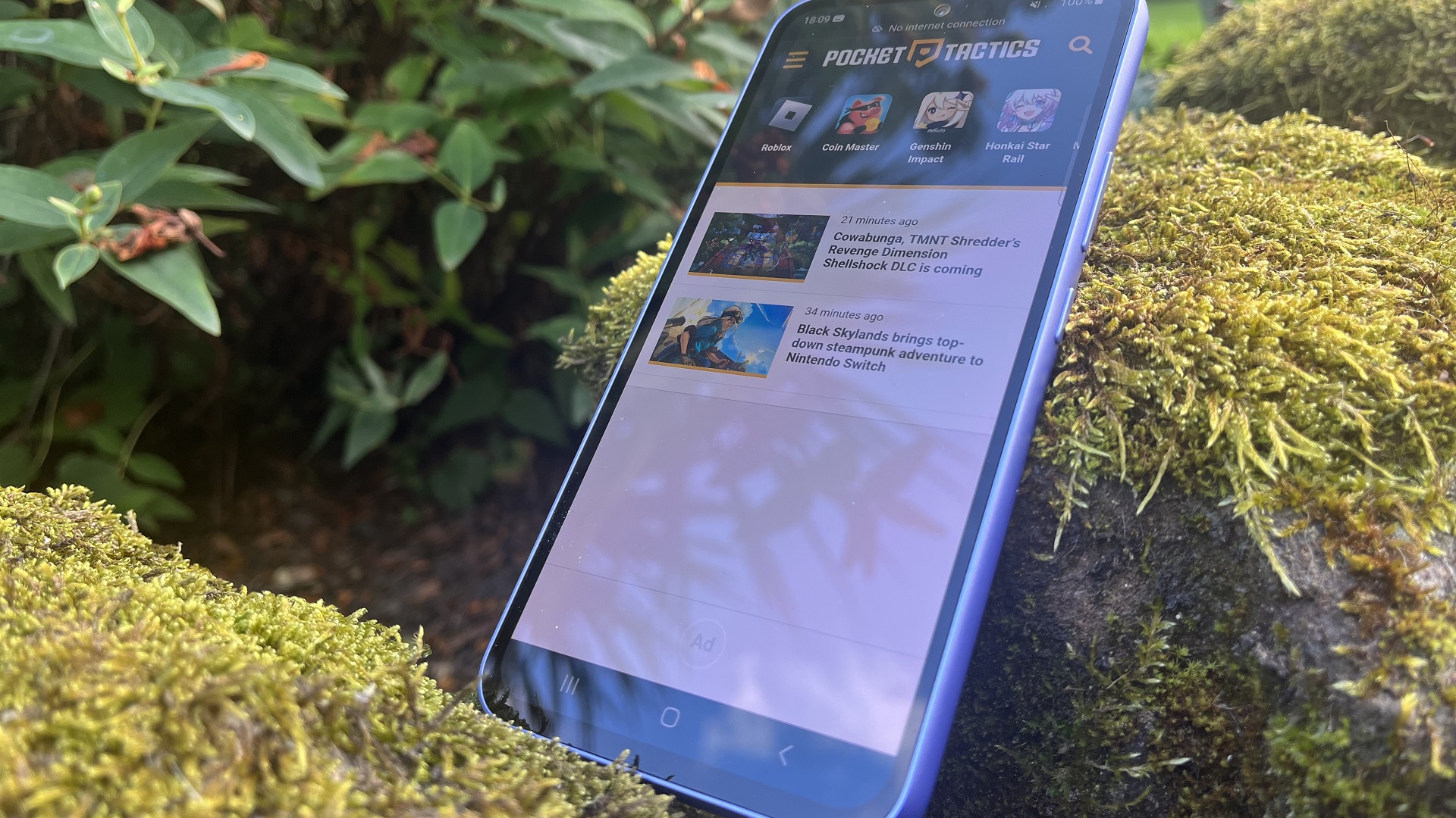 Samsung A54 レビュー - 苔むした岩の上に置かれた携帯電話と、画面に Pocket Tactics の Web ページが表示されている